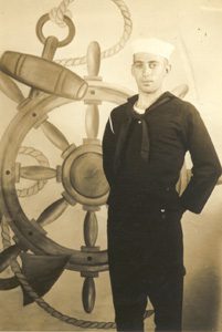 Sailor in uniform beside ship's wheel backdrop